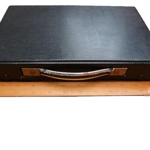 Peut inclure: Une mallette de transport rectangulaire noire avec une surface textur&eacute;e et une poign&eacute;e en m&eacute;tal argent&eacute;. Une r&egrave;gle en bois est plac&eacute;e devant la mallette, indiquant que la largeur de l'objet est d'environ 30,5 cm d'une extr&eacute;mit&eacute; &agrave; l'autre.