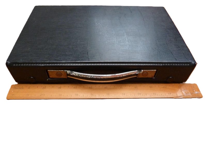 Peut inclure: Une mallette de transport rectangulaire noire avec une surface textur&eacute;e et une poign&eacute;e en m&eacute;tal argent&eacute;. Une r&egrave;gle en bois est plac&eacute;e devant la mallette, indiquant que la largeur de l'objet est d'environ 30,5 cm d'une extr&eacute;mit&eacute; &agrave; l'autre.