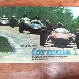 May include: A Waddingtons Formula 1 board game box cover featuring a color photograph of three vintage open-wheel race cars driving on a track. The text reads Waddingtons formula 1 an action packed motor racing game for 2 to 6 players.