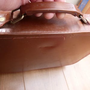 May include: A brown briefcase with a smooth, structured surface and a sturdy top handle. The handle is attached to the main body with metallic hardware, including small rivets and hinges, featuring visible stitching along the top edge.