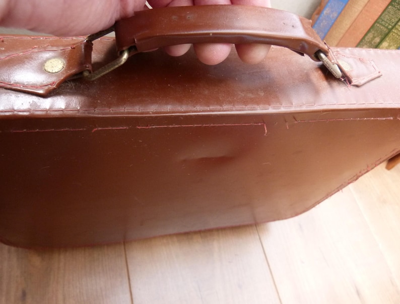 May include: A brown briefcase with a smooth, structured surface and a sturdy top handle. The handle is attached to the main body with metallic hardware, including small rivets and hinges, featuring visible stitching along the top edge.