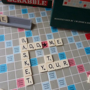 Puede incluir: Un primer plano de un tablero de Scrabble con fichas que deletrean la palabra "ADDS" y "YOUR" en un tablero gris y blanco con casillas rojas y azules. Las fichas son blancas con letras y n&uacute;meros negros.