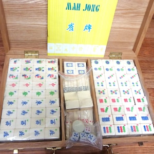 May include: A wooden Mah Jong game set in a hinged case with a yellow instruction booklet featuring the text MAH JONG. The set includes white tiles with colourful engraved symbols, a plastic bag containing white dice, and blank tiles.