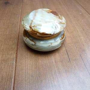 May include: A round, lidded trinket box crafted from polished green and white banded onyx stone. The container features a smooth, domed lid with natural brown veining and a metallic gold-toned rim connecting the base to the lid.