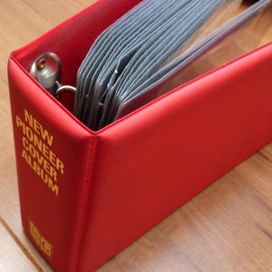 May include: A red photo album with the text "NEW PIONEER COVER ALBUM" in gold lettering. The album is open, revealing clear plastic sleeves filled with photos. The album has a metal ring binder.