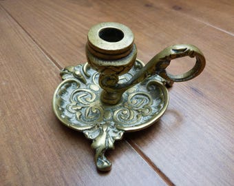 Puede incluir: Un candelabro de latón con un patrón floral en relieve en la base circular y un asa curva. La superficie metálica muestra una pátina envejecida y desgastada con detalles intrincados de volutas en toda la pieza.
