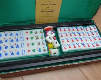 May include: A green Mah Jongg game set in a brown leather case. The case is open, revealing the tiles and a guide to the game. The tiles are white with red, blue, and green markings. The guide is titled "Mah-Jongg" and has a small logo that says "Jackpot".