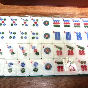 May include: A set of white rectangular mahjong tiles arranged in a grid inside a green tray, wrapped in clear plastic. Each tile features colorful circular patterns and red numbers, including ones, twos, threes, fours, fives, sixes, sevens, eights, and nines.