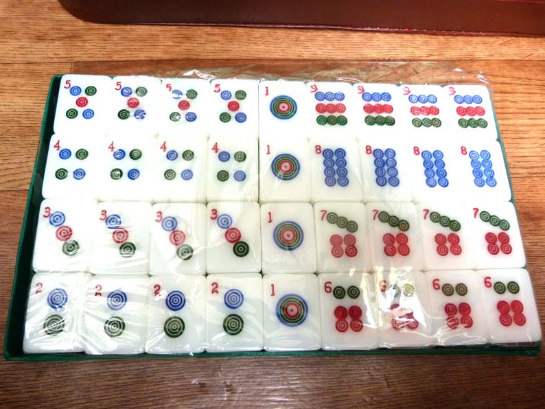 May include: A set of white rectangular mahjong tiles arranged in a grid inside a green tray, wrapped in clear plastic. Each tile features colorful circular patterns and red numbers, including ones, twos, threes, fours, fives, sixes, sevens, eights, and nines.