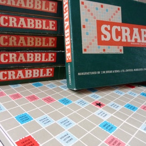 Puede incluir: Una pila de cinco cajas verdes y rojas con la palabra "SCRABBLE" impresa en cada caja. La caja superior est&aacute; abierta y muestra un tablero de juego verde con una estrella roja en el centro. El tablero de juego est&aacute; etiquetado con "DOUBLE LETTER SCORE", "TRIPLE LETTER SCORE", "DOUBLE WORD SCORE" y "TRIPLE WORD SCORE".