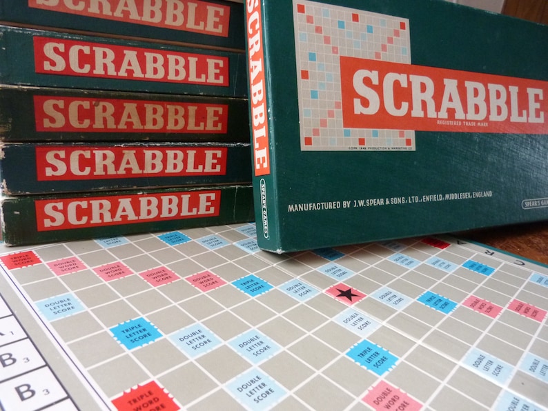 Puede incluir: Una pila de cinco cajas verdes y rojas con la palabra "SCRABBLE" impresa en cada caja. La caja superior est&aacute; abierta y muestra un tablero de juego verde con una estrella roja en el centro. El tablero de juego est&aacute; etiquetado con "DOUBLE LETTER SCORE", "TRIPLE LETTER SCORE", "DOUBLE WORD SCORE" y "TRIPLE WORD SCORE".