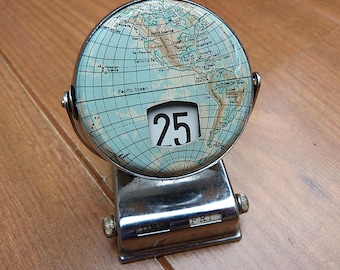 May include: A vintage-style desk calendar featuring a circular globe map face displaying North and South America. The center shows the number 25, while the chrome-finished base includes small manual windows displaying the month DEC and day FRI.