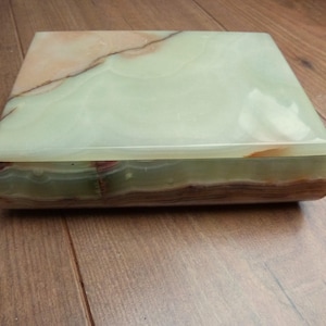 May include: A rectangular, lidded box made of polished onyx. The box features a light green top with brown and tan veining. The sides are a darker green and brown. The box is sitting on a wooden surface.