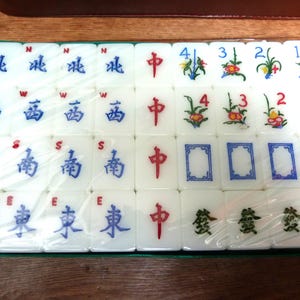 May include: A set of white Mahjong tiles arranged in a rectangular grid within a green tray, covered by clear plastic. The tiles feature blue directional characters, red central characters, green floral designs with numbers, and green symbols.