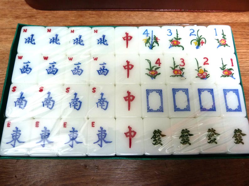 May include: A set of white Mahjong tiles arranged in a rectangular grid within a green tray, covered by clear plastic. The tiles feature blue directional characters, red central characters, green floral designs with numbers, and green symbols.