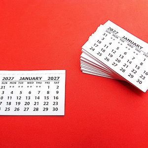 以下が含まれることがあります： 2027年1月の白い紙製カレンダーシートの束と、2027年1月のカレンダーシートが1枚。各シートには、日曜日から土曜日までの曜日を示す黒い印刷文字と数字が記載されています。