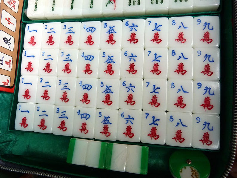 May include: A set of white Mahjong tiles arranged in a grid within a green case. Each tile features blue numbers one through nine at the top and red Chinese characters below. Four plain white and green tiles are placed horizontally at the bottom.