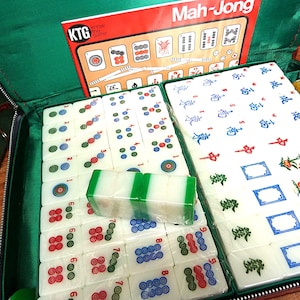 May include: A Mah-Jong game set in a green zippered case, featuring white tiles with colorful symbols, numbers, and characters. The set includes a KTG Know the Game instruction booklet, stacks of tiles, and red, green, and yellow plastic chips.