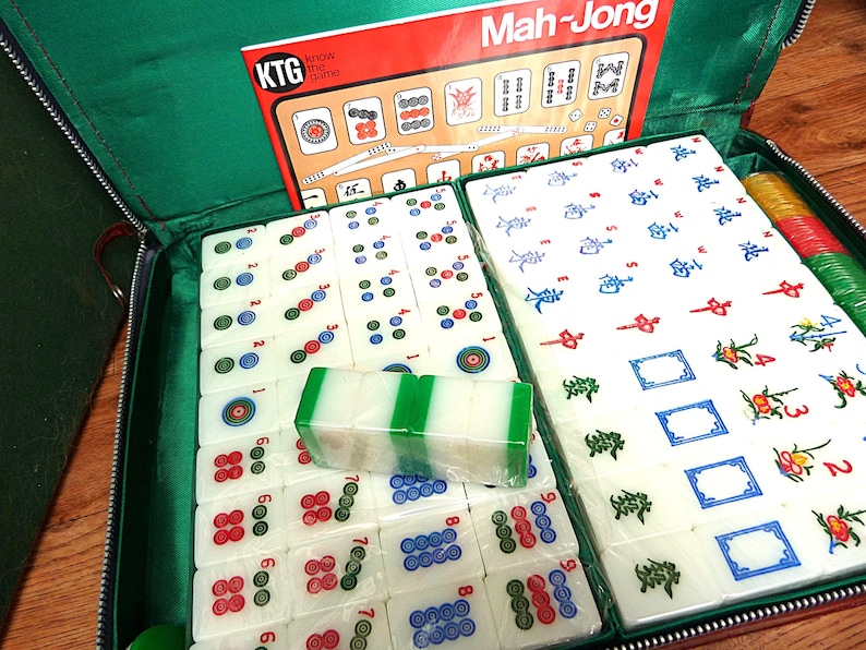 May include: A Mah-Jong game set in a green zippered case, featuring white tiles with colorful symbols, numbers, and characters. The set includes a KTG Know the Game instruction booklet, stacks of tiles, and red, green, and yellow plastic chips.