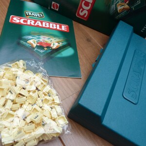 May include: A green travel edition Scrabble game box with the word "Scrabble" printed in white on the front. The box is closed and the lid is slightly ajar. The box is sitting on a wooden surface. A bag of letter tiles is in the foreground.