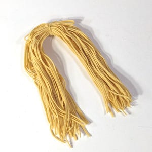 May include: A bundle of yellow shoelaces tied together at the top. The laces are a light, muted yellow color and appear to be made of a woven material. The laces are arranged in a cascading pattern against a white background.
