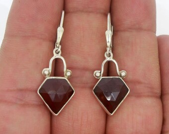 Op de afbeelding: Een paar zilveren oorhangers met een rode edelsteen in de vorm van een diamant. De oorbellen hebben een eenvoudig ontwerp met een kleine lus aan de bovenkant en een kleine bol aan elke kant van de edelsteen.
