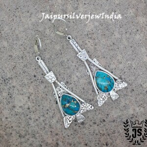Könnte beinhalten: Ein Paar silberfarbene Ohrringe mit einem türkisfarbenen Stein in einem dekorativen Rahmen. Die Ohrringe haben einen Klappverschluss und werden auf einer grauen Oberfläche präsentiert. Im Hintergrund ist der Text "JaipursilverjewIndia" zu sehen.