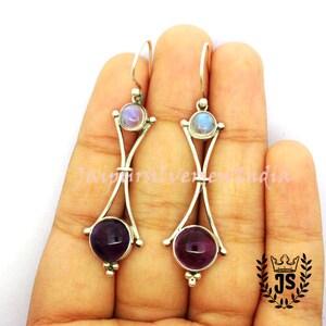 May include: A pair of silver dangle earrings with amethyst and moonstone gemstones. The earrings feature a delicate, curved design with a small silver ball at the bottom.