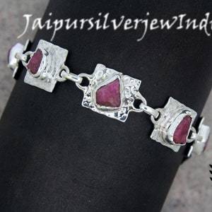 May include: Silver bracelet with four square-shaped charms, each featuring a pink gemstone. The bracelet is on a black surface.
