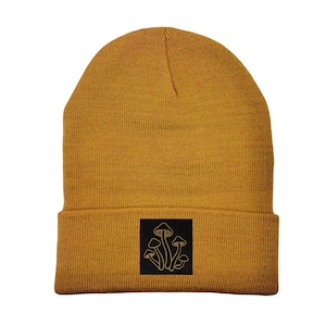 May include: A mustard yellow beanie with a black patch featuring a line drawing of golden mushrooms.