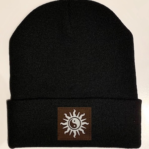 May include: Black beanie with a brown patch featuring a white yin yang symbol inside a sun design.