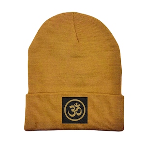 Puede incluir: Un gorro de punto amarillo mostaza con un parche cuadrado negro que presenta un símbolo Om dorado.