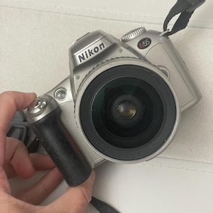 Könnte beinhalten: Eine silberne Nikon N55 Filmkamera mit einem schwarzen Objektiv und einem schwarzen Trageriemen.