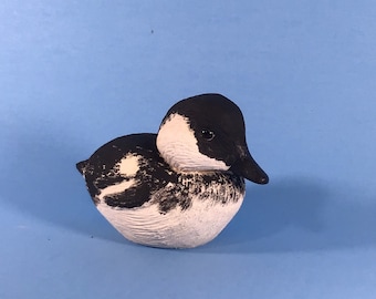 Puede incluir: Una figurita de cerámica pintada a mano de un pato blanco y negro con un pequeño pico negro. El pato está sentado sobre una superficie azul claro.