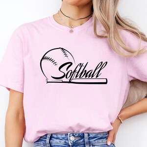K&ouml;nnte beinhalten: Ein hellrosa T-Shirt mit kurzen &Auml;rmeln und einem schwarzen Grafikdruck auf der Brust. Das Design zeigt einen Softball und einen Schl&auml;ger, wobei das Wort Softball in einer kursiven Schriftart in der Mitte abgebildet ist.
