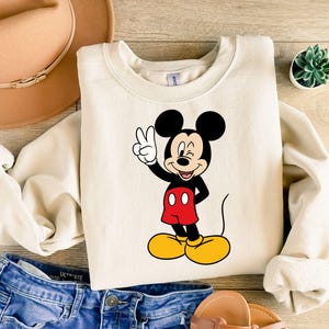 Pu&ograve; includere: Una felpa girocollo color crema con una stampa grafica di Topolino che fa l'occhiolino e il segno di vittoria con una mano. Il personaggio &egrave; raffigurato nei suoi classici colori nero, rosso e giallo, posizionato al centro del capo.
