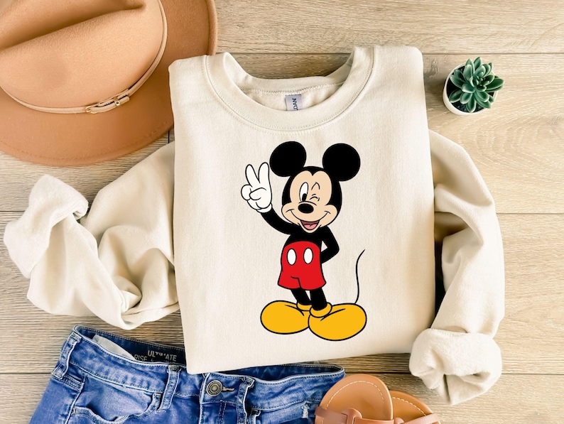 Pu&ograve; includere: Una felpa girocollo color crema con una stampa grafica di Topolino che fa l'occhiolino e il segno di vittoria con una mano. Il personaggio &egrave; raffigurato nei suoi classici colori nero, rosso e giallo, posizionato al centro del capo.