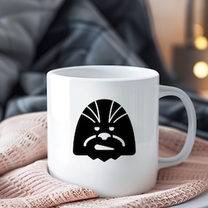 May include: A white ceramic mug featuring a black graphic illustration of a stylized Wookiee character face on the front. The mug has a smooth, glossy finish with a standard handle on the right side and a simple, cylindrical shape for holding beverages.