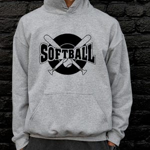 K&ouml;nnte beinhalten: Ein hellgrauer Kapuzenpullover mit einem schwarzen Grafikdruck auf der Brust. Das Design zeigt das Wort SOFTBALL in fetten Gro&szlig;buchstaben, &uuml;berlagert von zwei gekreuzten Baseballschl&auml;gern und einem Softball in einem soliden schwarzen Kreis.