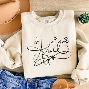 May include: A cream-colored crewneck sweatshirt featuring a black graphic design of the name Ariel written in a stylized, cursive script with small circular accents above the lettering, displayed alongside a tan hat and blue denim jeans.