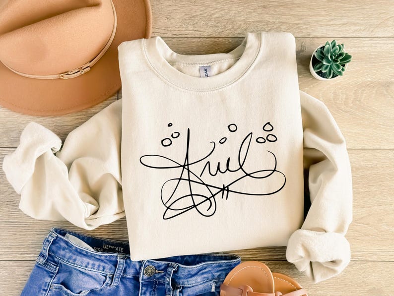 May include: A cream-colored crewneck sweatshirt featuring a black graphic design of the name Ariel written in a stylized, cursive script with small circular accents above the lettering, displayed alongside a tan hat and blue denim jeans.