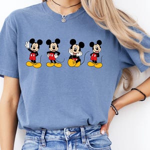 Pu&ograve; includere: Una t-shirt blu a maniche corte con una stampa grafica di quattro diverse pose di Topolino. Ogni illustrazione del personaggio mostra Topolino con orecchie nere, pantaloncini rossi e scarpe gialle, disposti orizzontalmente sul petto.
