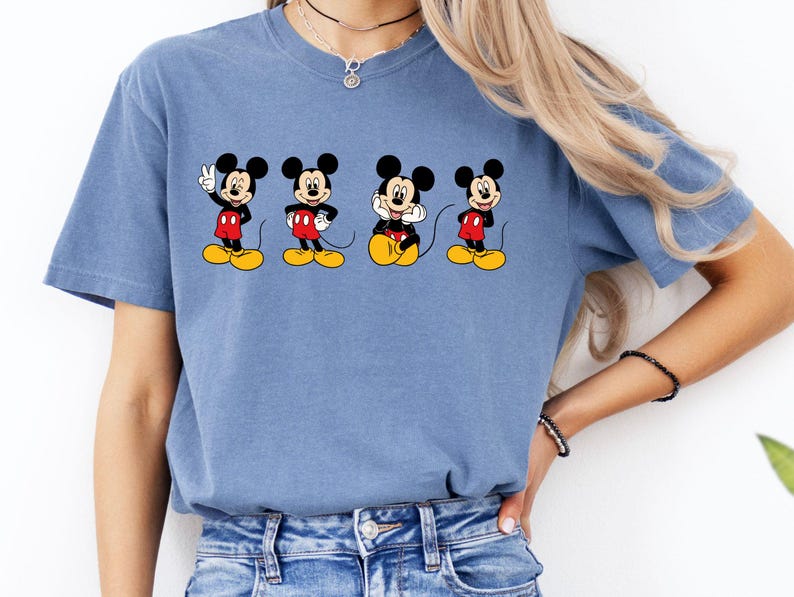 Pu&ograve; includere: Una t-shirt blu a maniche corte con una stampa grafica di quattro diverse pose di Topolino. Ogni illustrazione del personaggio mostra Topolino con orecchie nere, pantaloncini rossi e scarpe gialle, disposti orizzontalmente sul petto.
