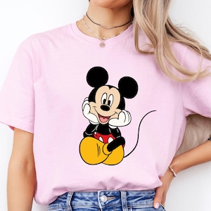 Pu&ograve; includere: Una t-shirt a maniche corte color rosa chiaro con una stampa centrale di Topolino seduto con le mani appoggiate sulle guance. Il personaggio &egrave; raffigurato con orecchie nere, viso color crema, maglietta rossa e grandi scarpe gialle rotonde.