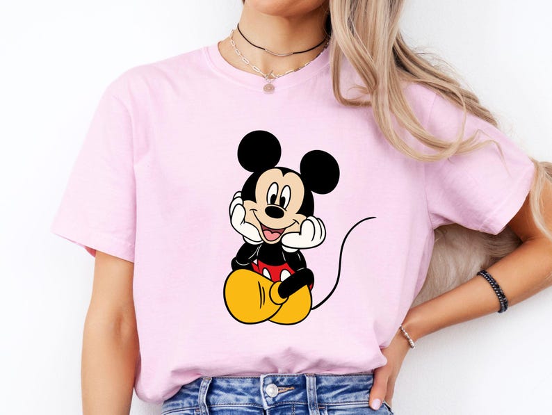 Pu&ograve; includere: Una t-shirt a maniche corte color rosa chiaro con una stampa centrale di Topolino seduto con le mani appoggiate sulle guance. Il personaggio &egrave; raffigurato con orecchie nere, viso color crema, maglietta rossa e grandi scarpe gialle rotonde.