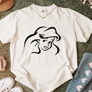 May include: A cream-colored short-sleeve t-shirt featuring a black line art graphic of a female character with long flowing hair centered on the chest. The shirt has a crew neckline and a visible brand tag at the inner collar area.