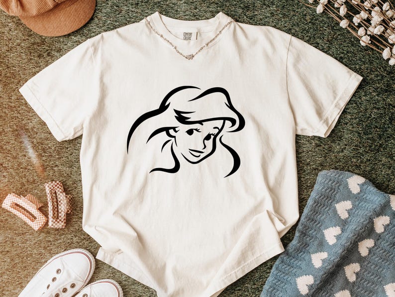 May include: A cream-colored short-sleeve t-shirt featuring a black line art graphic of a female character with long flowing hair centered on the chest. The shirt has a crew neckline and a visible brand tag at the inner collar area.