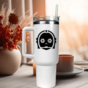May include: A white stainless steel tumbler with a handle, a clear lid, and a clear straw. The front of the tumbler features a black graphic of a robotic head with circular eyes and a segmented face design, centered on the upper portion of the body.