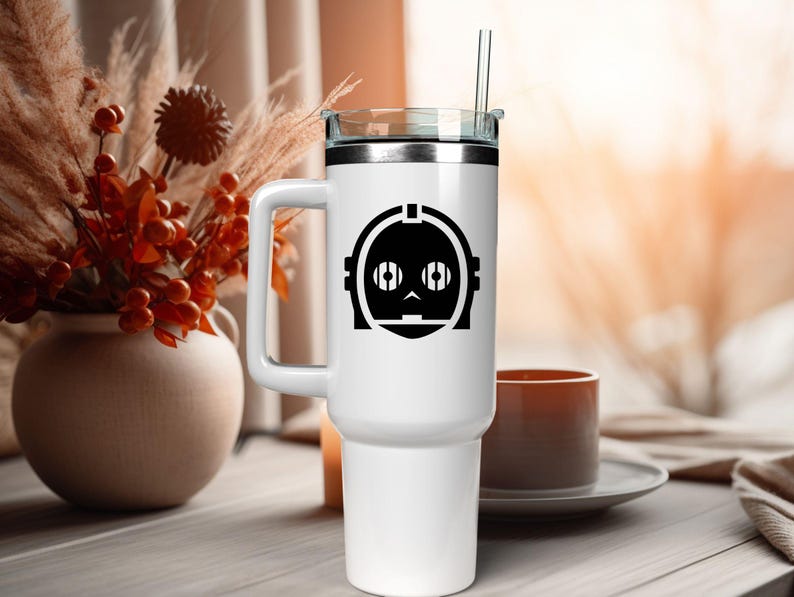 May include: A white stainless steel tumbler with a handle, a clear lid, and a clear straw. The front of the tumbler features a black graphic of a robotic head with circular eyes and a segmented face design, centered on the upper portion of the body.