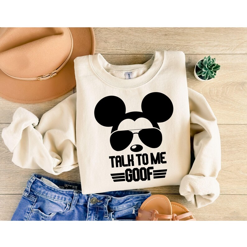 May include: A cream-colored crewneck sweatshirt featuring a black graphic of a mouse head wearing sunglasses with the text "TALK TO ME GOOF" printed underneath. The sweatshirt is displayed alongside a tan hat, blue denim jeans, and sandals.